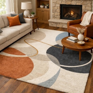 May include: A large area rug with an abstract design. The rug features overlapping circles in shades of rust, gray, beige, and blue, with a gold-colored curved line. The rug is placed on a hardwood floor in a living room setting.