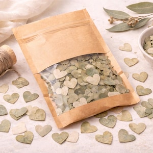 May include: A kraft paper bag with a clear window filled with heart-shaped eucalyptus leaves. Scattered around are more heart-shaped leaves in shades of green and cream. A small bowl of leaves and a spool of twine are also present.