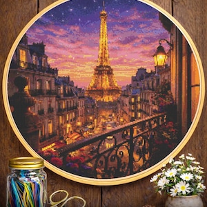 Pode incluir: Uma bordado de ponto cruz da Torre Eiffel ao pôr do sol, emoldurado em um aro dourado. A cena inclui edifícios, uma varanda e um céu estrelado. Um pote de fios coloridos, tesouras e um pequeno vaso de flores estão em primeiro plano.