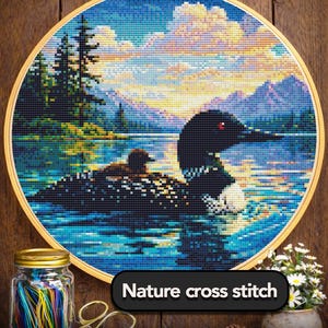 May include: A cross-stitch embroidery of a loon on a lake, set in a wooden hoop. The scene includes a sunset, mountains, and trees. The text "Nature cross stitch" is displayed on a black banner. A jar of colorful threads and scissors are in the foreground.