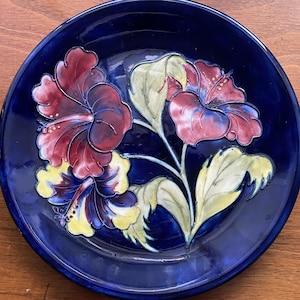 May include: A round ceramic plate with a deep cobalt blue glaze featuring a raised, embossed floral design. The central motif displays three large hibiscus-style flowers in shades of dark red, burgundy, and yellow, accompanied by light green leaves.