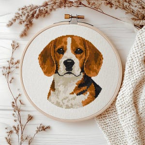 May include: A cross-stitch embroidery of a beagle dog's head and shoulders, set in a wooden hoop. The dog has brown and white fur, with black accents. The background is white, and the hoop is surrounded by dried plants and a cream-colored knitted blanket.