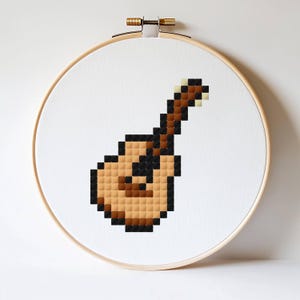 Puede incluir: Un diseño de punto de cruz de una guitarra pixelada en tonos marrones, negros y beige, exhibido dentro de un aro de bordado de madera. La guitarra está centrada en un fondo de tela blanca.