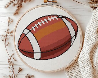 Könnte beinhalten: Ein Football-Design im Kreuzstich in einem hölzernen Stickrahmen. Der Football ist braun mit weißen Streifen und Nähten. Der Hintergrund ist weißer Stoff. Der Rahmen befindet sich auf einer weißen Holzoberfläche mit getrockneten Pflanzen und einer cremefarbenen Strickdecke.
