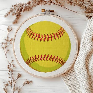 May include: A cross-stitch embroidery hoop featuring a yellow softball with red stitching details. The embroidery is set against a white fabric background, framed by a wooden hoop. The design is a sports-themed craft.