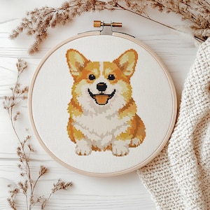 May include: A cross-stitch embroidery of a smiling Corgi dog in a wooden hoop. The dog is depicted with orange, white, and black thread. The background is white fabric. The hoop is set against a white wooden surface with dried plants and a cream-colored knitted blanket.