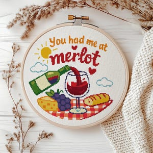 Könnte beinhalten: Kreuzstich-Stickerei in einem Holzreifen mit dem Text "You had me at merlot". Das Design zeigt eine Weinflasche, ein Glas Rotwein, Käse, Trauben und ein Baguette auf einer rot-weiß karierten Picknickdecke.