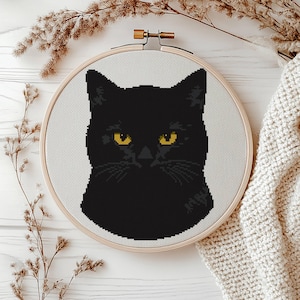 May include: A circular wooden embroidery hoop containing a cross-stitch pattern of a black cat head with bright yellow eyes. The cat is centered on a white fabric background, and the hoop features a metal tightening screw at the top center.