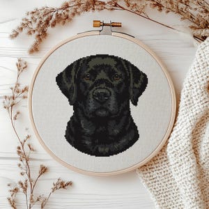 May include: A cross-stitch portrait of a black Labrador Retriever, framed in a wooden embroidery hoop. The dog's image features brown eyes and a black coat. The hoop is presented on a white surface, with dried flowers and a cream-colored knitted blanket in the background.