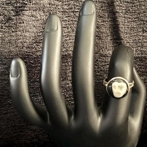 May include: A silver ring with a black and white oval-shaped design. The ring is displayed on a black hand-shaped display. The design appears to be a tooth.