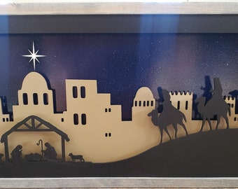 May include: A framed wooden Christmas scene featuring a silhouette of a town, a nativity scene, and the three wise men on camels. The background is a starry night sky. The frame is silver.