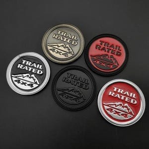 May include: A collection of five circular Trail Rated 4x4 badges in various colors. The badges feature a mountain range graphic and are in silver, black, bronze, red, and black. The text "TRAIL RATED" and "4X4" are prominently displayed.