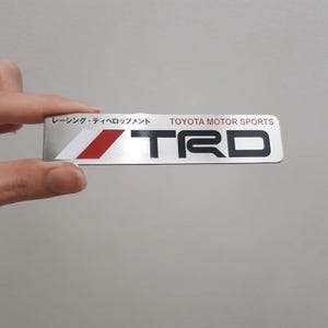 May include: A rectangular silver TRD emblem with the text "TOYOTA MOTOR SPORTS" and Japanese characters. The emblem features a red and white diagonal stripe. The letters "TRD" are in bold black.