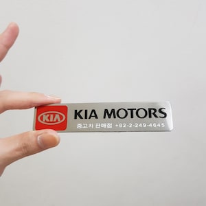 Peut inclure: Un emblème rectangulaire argenté Kia Motors avec le logo Kia dans un ovale rouge à gauche. Le texte "KIA MOTORS" est en lettres noires en gras. En dessous, du texte coréen et un numéro de téléphone sont imprimés.