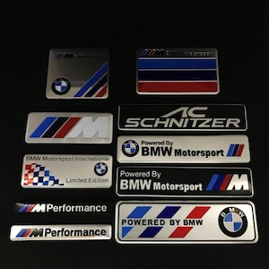 Peut inclure: Collection d'emblèmes et badges automobiles, avec le logo BMW et divers designs Motorsport. Les badges sont argentés, noirs, rouges et bleus, avec des textes tels que "M Performance" et "AC Schnitzer".