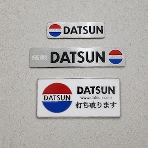 May include: Three Datsun car emblems. The top emblem is rectangular with the word "Datsun" in black text. The middle emblem has Japanese characters and the word "Datsun". The bottom emblem has the website address and Japanese text.