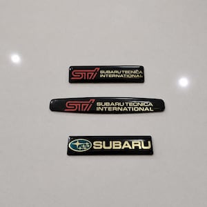 May include: Three black and gold Subaru car emblems. Two emblems have the STI logo with "SUBARU TECNICA INTERNATIONAL" text. The third emblem has the Subaru logo with the word "SUBARU".