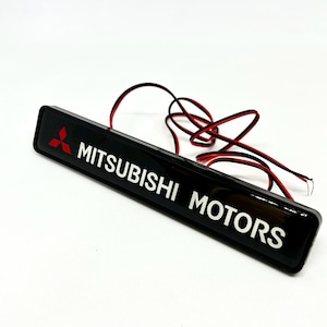 May include: A rectangular black emblem featuring the red Mitsubishi three-diamond logo and the white text MITSUBISHI MOTORS. The glossy surface is attached to two thin red and black electrical wires extending from the back of the unit.