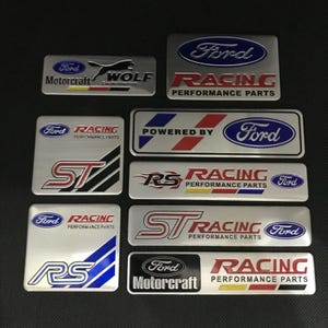 May include: A selection of Ford Racing and Motorcraft emblem badges. The badges display the Ford logo and text like "RACING PERFORMANCE PARTS", "ST", and "RS". The colour scheme includes silver, red, blue, and black. Various shapes and sizes are shown.