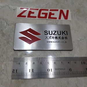 May include: Two metal Suzuki emblems and a Zegen sticker. The Suzuki emblem is rectangular with a red logo and the word SUZUKI in black. The Japanese text and website address are also visible. A stainless steel ruler is below the emblems.