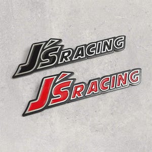 Könnte beinhalten: Zwei Autoembleme mit dem Text "JS RACING". Eines ist schwarz mit weißem Rand, das andere rot mit weißem Rand. Beide liegen auf einer hellgrauen Oberfläche.