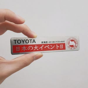 Puede incluir: Una insignia rectangular de Toyota con texto japonés y un número de teléfono. La insignia tiene un fondo plateado con texto negro "TOYOTA" y una sección roja con caracteres japoneses. Un pequeño logotipo está a la derecha.