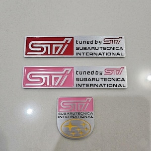 Peut inclure: Trois emblèmes Subaru Tecnica International (STI). Deux emblèmes rectangulaires présentent le logo STI en rouge et rose, avec le texte "tuned by STI" et "SUBARU TECNICA INTERNATIONAL". Le troisième emblème est rose et argenté avec un design d'étoile dorée.