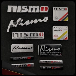 Könnte beinhalten: Eine Sammlung von Nismo-Autoemblemen in verschiedenen Formen und Größen. Die Embleme zeigen das Nismo-Logo und den Text, einige enthalten den Text 'Nissan Motorsports International'. Farben sind Silber, Schwarz, Rot und Weiß.
