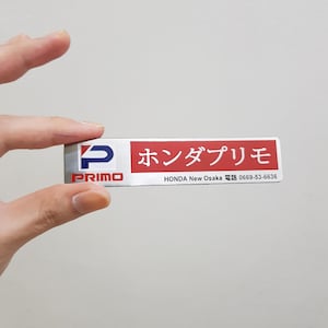 May include: A rectangular sticker with a red and white background. The sticker has the word "PRIMO" in white text with a blue "P" logo on the left side. The right side has Japanese text and the words "HONDA New Osaka" and a phone number.