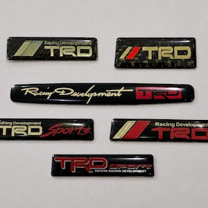Op de afbeelding: Een verzameling van zes rechthoekige auto-emblemen met het TRD-logo. De badges tonen verschillende kleurencombinaties, waaronder zwart, rood, goud en koolstofvezelpatronen met de tekst Racing Development en TRD Sport.