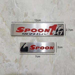 Puede incluir: Dos emblemas rectangulares plateados de Spoon. El emblema más grande mide 12 cm de largo y presenta texto rojo que dice "Spoon" y "Tune Up" con caracteres japoneses. El emblema más pequeño mide 7 cm de largo con texto "Spoon" en rojo.
