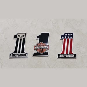 Può includere: Tre emblemi Harley-Davidson numero uno. Uno presenta un teschio, un altro il logo Harley-Davidson e il terzo ha un design rosso, bianco e blu. Ogni emblema ha il nome Harley-Davidson.