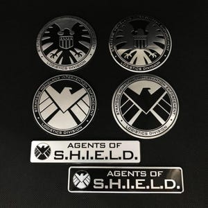 May include: A selection of silver and black metal emblems and badges. The round emblems display a stylized eagle and the text "Strategic Homeland Intervention Enforcement Logistics Division." Rectangular badges read "Agents of S.H.I.E.L.D."