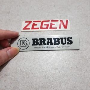 May include: Two rectangular metal car emblems. The top emblem reads "ZEGEN" in red letters on a white background. The bottom emblem reads "BRABUS" in black letters on a silver background, with the text "Brabus for Mercedes-Benz product".