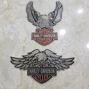 Könnte beinhalten: Zwei Harley-Davidson-Embleme. Das obere Emblem zeigt einen silbernen Adler mit ausgebreiteten Flügeln über einem rechteckigen Logo mit den Worten "MOTOR HARLEY-DAVIDSON CYCLES" in Schwarz und Orange. Das untere Emblem hat ein ähnliches Design.