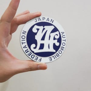 May include: A round, silver-colored emblem with the text "JAPAN AUTOMOBILE FEDERATION" in white. The emblem features a blue circle with a white stylized "JAF" logo in the center. The emblem is held by a hand.
