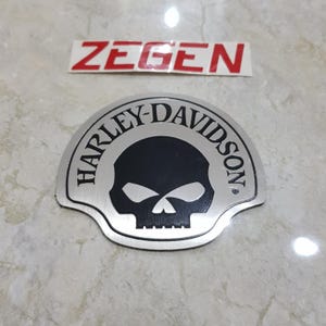 Puede incluir: Un emblema plateado de Harley-Davidson con un diseño de calavera negra. El emblema es semicircular con las palabras "HARLEY-DAVIDSON" en una fuente curva. Encima del emblema hay una pegatina roja y blanca con la palabra "ZEGEN".