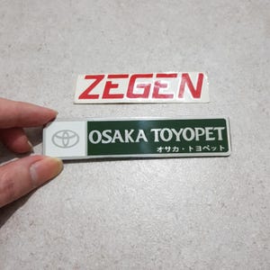 May include: Two rectangular car emblems. The top emblem has the word "ZEGEN" in red letters on a white background. The bottom emblem has the words "OSAKA TOYOPET" in white letters on a green background, with the Toyota logo and Japanese text.