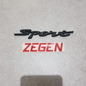 Puede incluir: Emblema negro de 'Sport' y una pegatina roja y blanca de 'Zegen'. El emblema de 'Sport' está en una fuente cursiva. La pegatina de 'Zegen' tiene texto rojo sobre fondo blanco. Los elementos están sobre una superficie gris.