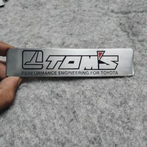 Puede incluir: Un emblema rectangular de metal plateado con el texto "TOM'S" en negro y rojo, y el texto "PERFORMANCE ENGINEERING FOR TOYOTA" en negro. El emblema está sostenido por una mano.