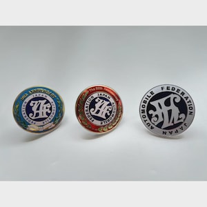 May include: Three round pins with the text "JAPAN AUTOMOBILE FEDERATION". One pin is blue with "20th ANNIVERSARY", another is red with "The 50th Anniversary", and the third is silver and black.