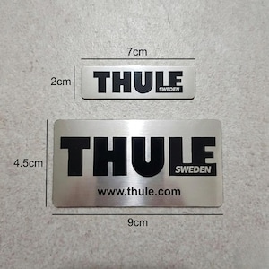 May include: Two silver rectangular Thule brand stickers. The larger sticker is 9 cm long and 4.5 cm wide, with the text "THULE SWEDEN" and "www.thule.com". The smaller sticker is 7 cm long and 2 cm wide, with the text "THULE SWEDEN".