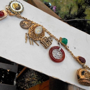 May include: A gold chain necklace with multiple charms, including a cameo, a lock, a green stone, and a red stone. The charms are attached to a gold chain with a lobster clasp.