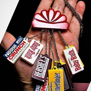 Puede incluir: Un collar original con un colgante rojo y blanco y dijes de barras de caramelo en miniatura. Los dijes representan barras "Nestle Crunch", "Baby Ruth", "Hershey's" y "Butterfinger". El collar está hecho de metal.
