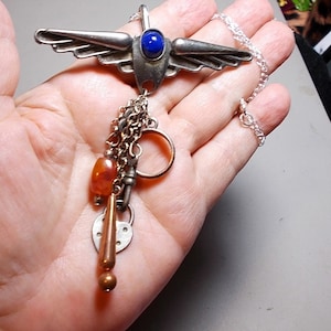 Puede incluir: Un collar de plata con un colgante alado con una piedra preciosa azul. El colgante tiene un dije con un corazón, una llave y una pequeña cuenta de ámbar.