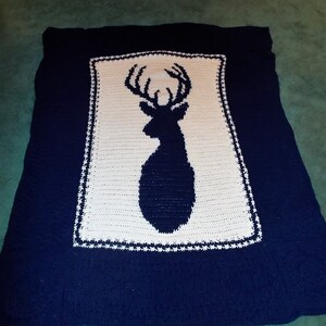 Puede incluir: Una manta de crochet azul marino con una silueta de ciervo de crochet blanco en el centro.