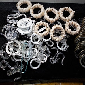 May include: An assortment of curtain hardware, including clear plastic hooks, white rings, fabric-covered rings, and metal rings. The items are arranged on a black surface, providing a contrast to the various colors and textures of the hardware.