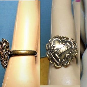 Puede incluir: Un anillo de tono dorado con un diseño en forma de corazón que presenta las palabras "PRO JESU" en una pancarta. El anillo se lleva en un dedo.