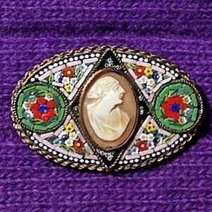 May include: A vintage brooch with a cameo of a woman's profile surrounded by colorful mosaic tiles. The brooch is oval shaped and has a gold tone metal frame.