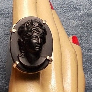 Könnte beinhalten: Ein schwarzer Kameenring mit einer goldenen Fassung. Die Kamee zeigt das Profil einer Frau mit einem Lorbeerkranz.